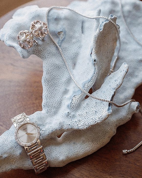 Wedding jewelry flatlay featuring a diamond tennis necklace, floral stud earrings and wristwatch with coral accent on a wood tabletop