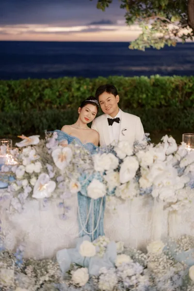 Couple portrait by candlelight, set against an ocean horizon and sunset sky, with a greenery hedge and overhanging tree branches behind.