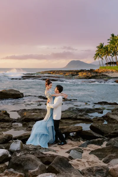 Couple portrait, a beach wedding photo with the groom in a tuxedo embracing his bride by waves on a rocky shore with palm trees under a sunset sky.