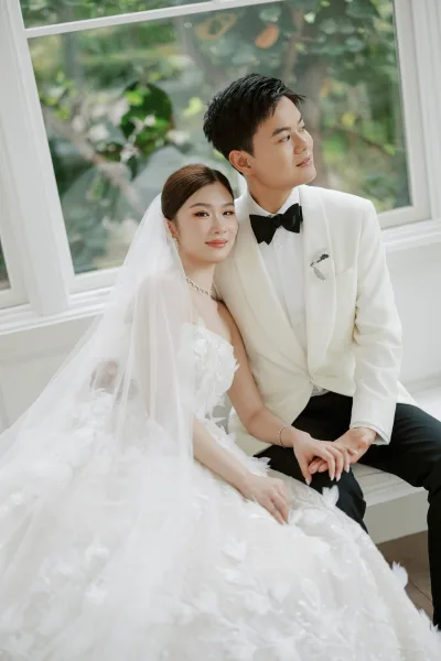 Couple portrait of bride and groom holding hands, her veil catching light by a window with garden greenery framing the scene.