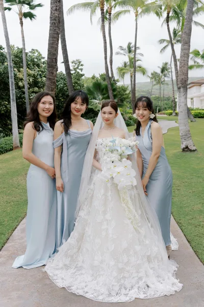 Bridal party portrait of a bride with bridesmaids in light blue satin dresses, the bride holding a cascading orchid bouquet by palm-lined gardens.