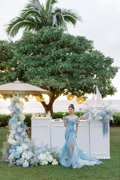 Bridal portrait in a blue wedding dress, the bride holds a cocktail glass beneath palm trees with the ocean and sky beyond.