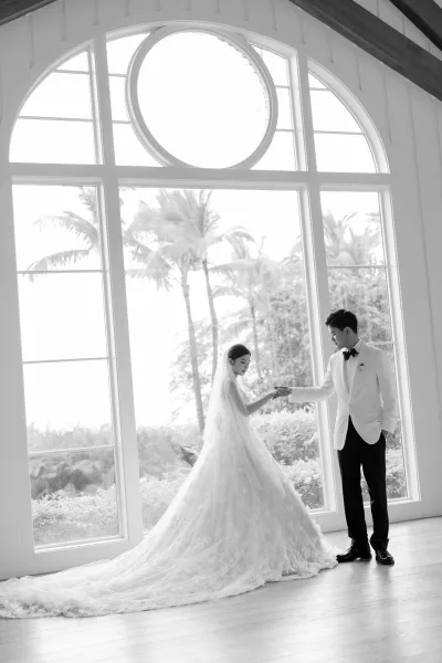 Couple portrait of the bride and groom holding hands beneath an arched window, her cathedral veil trailing as palm trees glow outside.