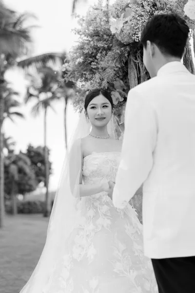 Ceremony moment as the wedding vows exchange unfolds beneath a floral arch, framed by palm trees on a sunlit lawn.