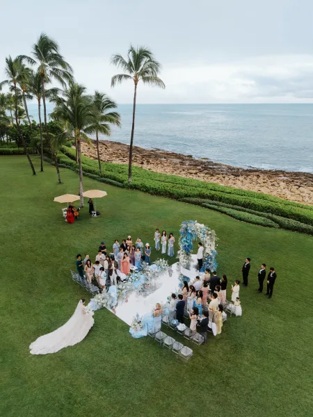 Ceremony processional as the bride walks down a mirrored aisle, ocean and palm trees beyond a rocky shore framing the sunlit lawn setting.