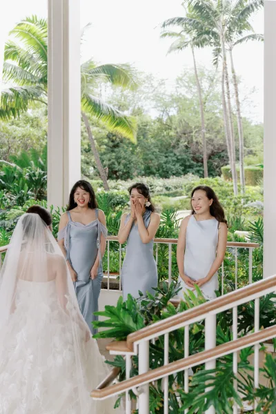 Bridesmaid first look during a bride reveal to bridesmaids, her long bridal veil catching light on a veranda with palm trees.