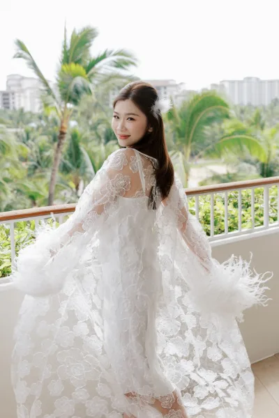 Bridal portrait of a bride on balcony in a sheer lace bridal robe, framed by palm trees and resort buildings under a bright sky.