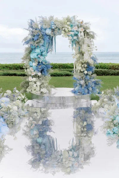 Wedding arch as a blue and white floral arch framing an ocean backdrop on a lawn, reflected in a mirror aisle.