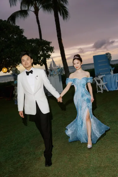 Couple portrait featuring a bride in blue gown holding hands with her partner on a palm-lined lawn by the ocean beneath a sunset sky.