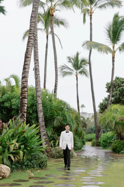 Groom portrait of a white dinner jacket groom on a stone path in a palm-lined tropical garden, his black bow tie crisp.