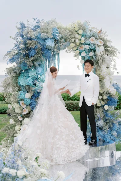 Wedding vows during an outdoor wedding ceremony as the couple stands beneath a floral arch, with the ocean and sky stretching behind on a lawn.