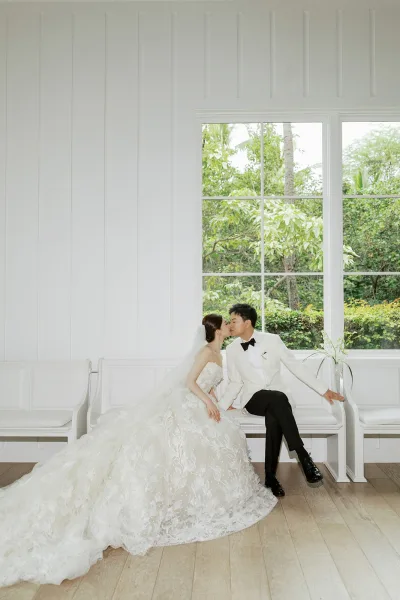 Wedding kiss portrait, a bench pose wedding photo of a bride with a long veil and groom seated on a pew by a large window with garden trees