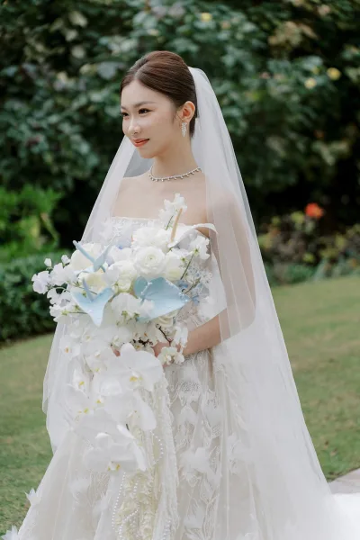 Bridal portrait of a bride with bouquet featuring cascading white orchids, set against lush garden greenery and neat lawn shrubs.