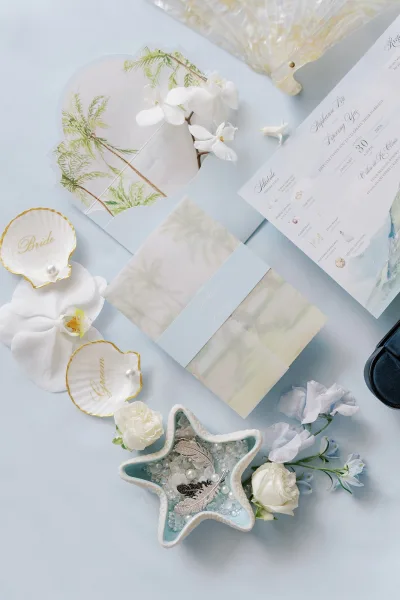 Wedding invitation suite styled as beach wedding invitations with seashells on a light blue surface, arranged with coordinating cards.