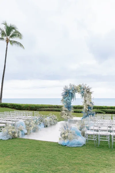 Ceremony setup with elegant outdoor ceremony decor, centered on a floral arch, facing the ocean with a lone palm tree over a manicured lawn.