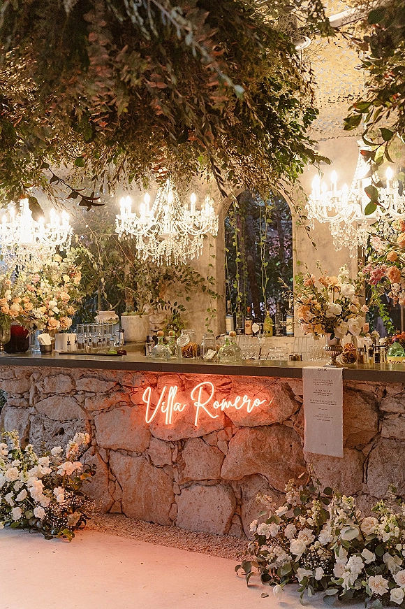Wedding bar decor with crystal chandeliers and hanging greenery above a stone bar featuring neon sign, menu, bottles, and candles