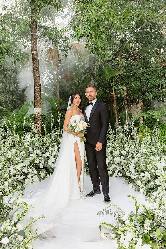 Couple portrait of bride holding a white bouquet beside groom in tuxedo, her cathedral veil trailing in a tropical garden aisle