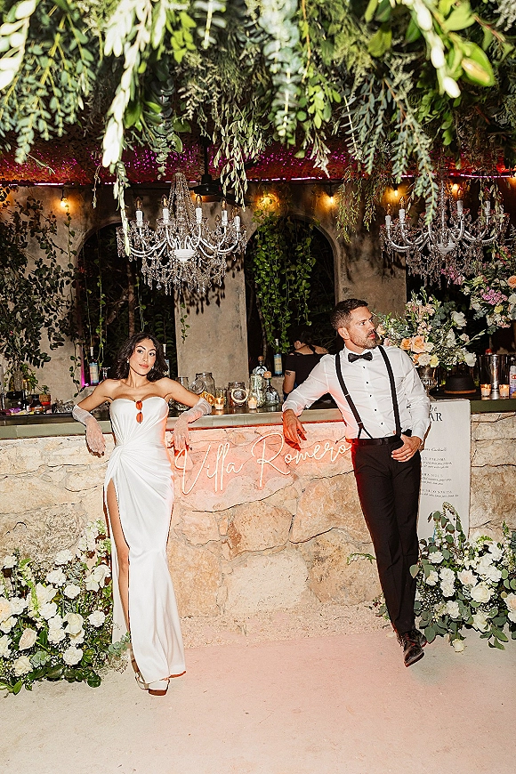 Couple portrait of bride and groom at bar, bride in strapless slit gown with opera gloves beside groom in bow tie and suspenders under chandeliers