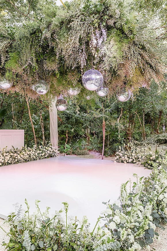 Wedding dance floor with silver disco balls hanging above a round white floor, framed by lush greenery, white florals, and draped backdrop