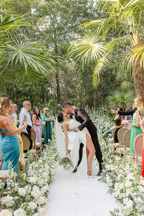 Wedding kiss as bride in veil holds bouquet and groom in tux dips her on a rose-petal aisle in a tropical garden setting