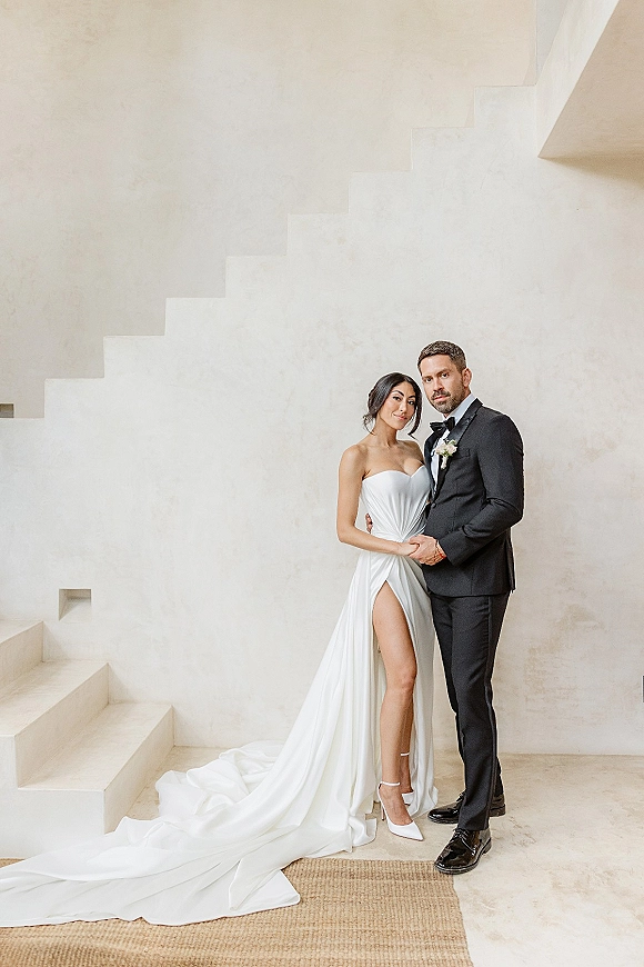 Couple portrait of bride and groom pose on a staircase by a white plaster wall, her strapless gown with thigh slit and train, his black tuxedo