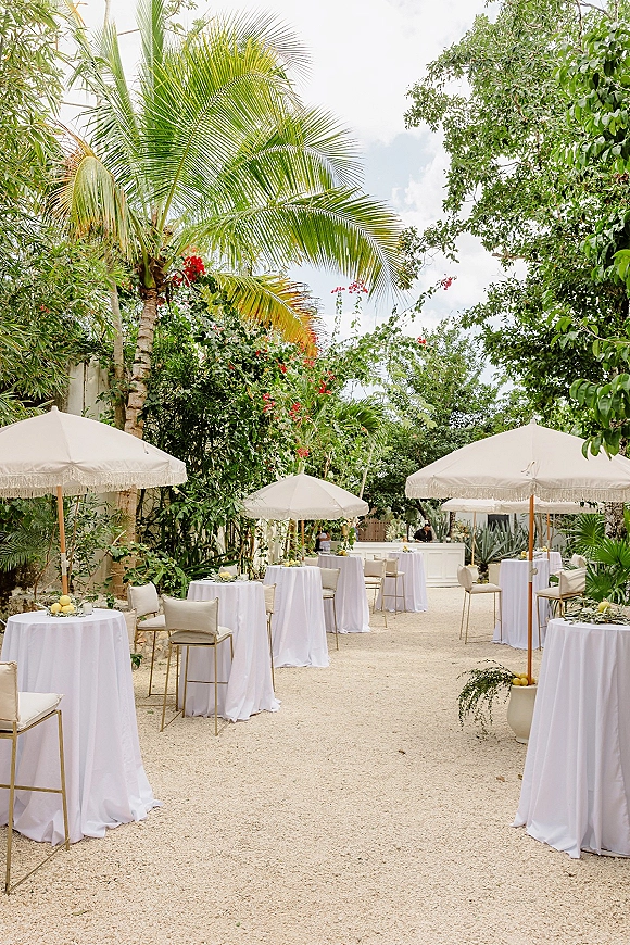 Outdoor cocktail reception with white linen high top cocktail tables under patio umbrellas, citrus centerpieces, and a bar in a tropical garden setting