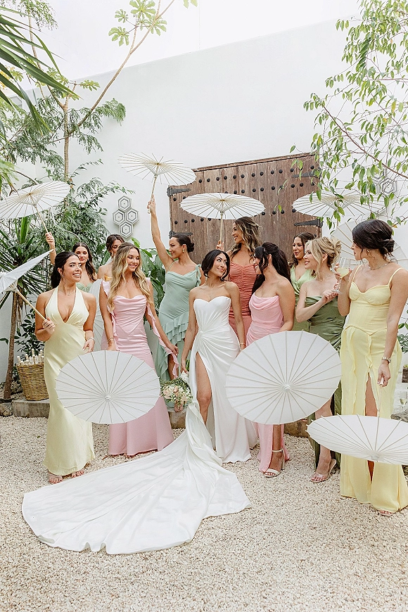 Bride with bridesmaids in pastel dresses holding paper parasols, bride in a white gown with train by rustic wooden doors in a courtyard