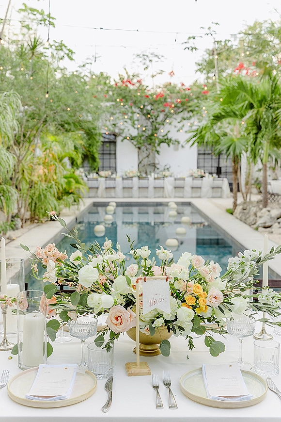Reception tablescape at a poolside wedding reception with gold compote roses, taper candles, menus and place cards on a long table under string lights
