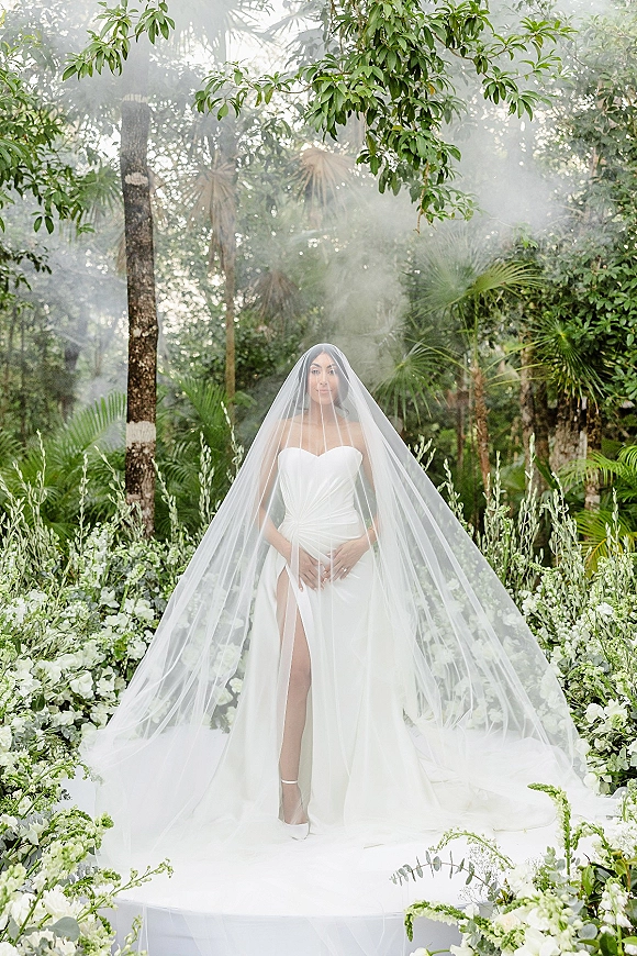 Bridal portrait of a cathedral veil bride in a strapless satin gown with thigh slit, standing on a misty garden aisle lined with white flowers