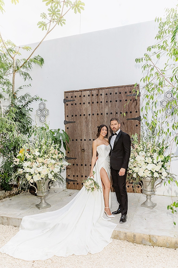 Couple portrait of bride and groom posing, bride in strapless long-train gown holding bouquet beside rustic wooden door courtyard backdrop