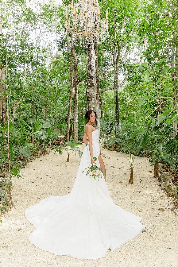 Bridal portrait of a bride in a strapless wedding dress with long train, holding a bouquet on a sandy forest path under crystal chandelier