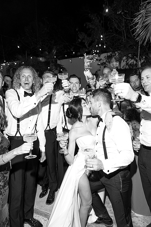 Wedding kiss as newlyweds champagne toast with raised coupe glasses, guests cheering under string lights on an outdoor patio at night