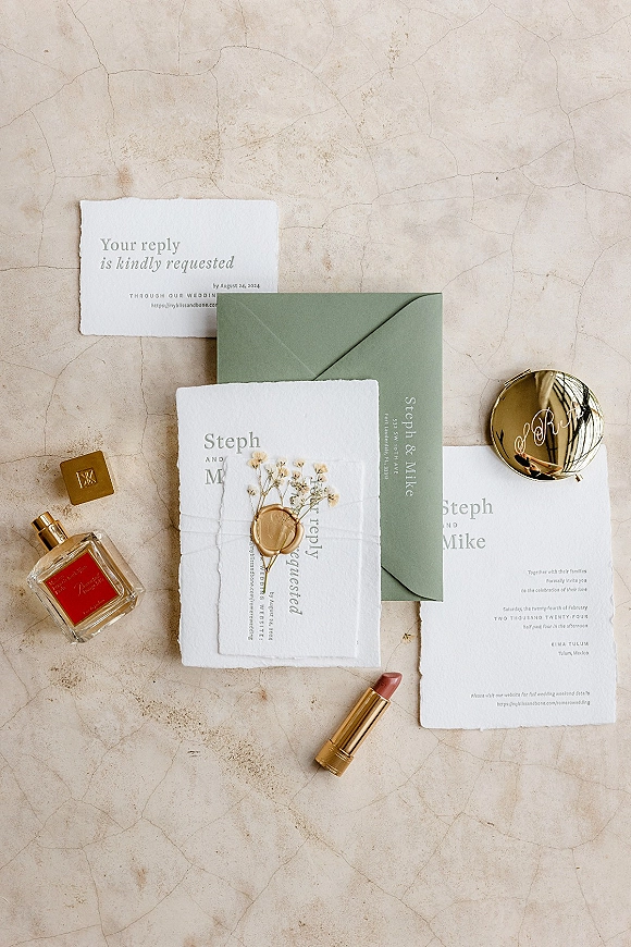 Invitation suite with deckled edge invitations and a sage green envelope, styled with wax seal on a textured stone surface flat lay