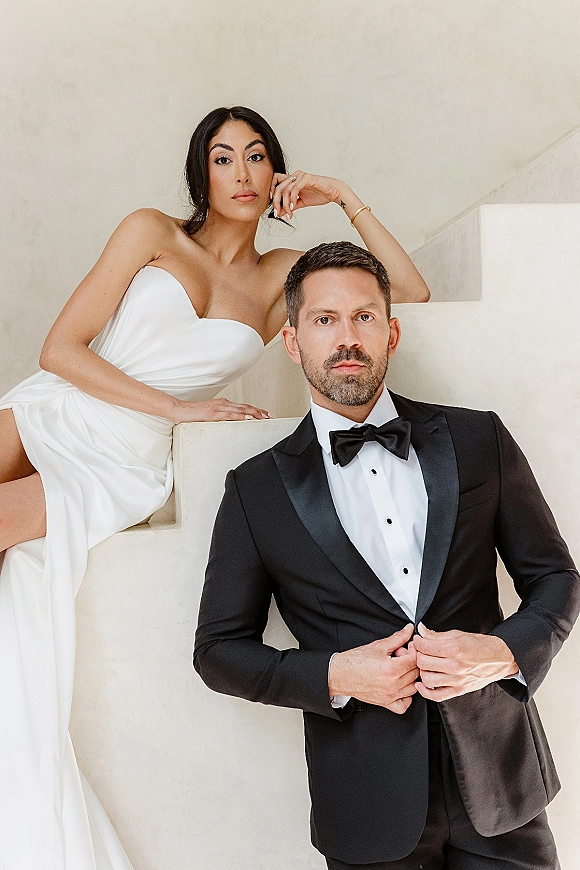 Couple portrait of bride in a strapless wedding dress and groom in a black tuxedo posing on a white staircase against a neutral wall