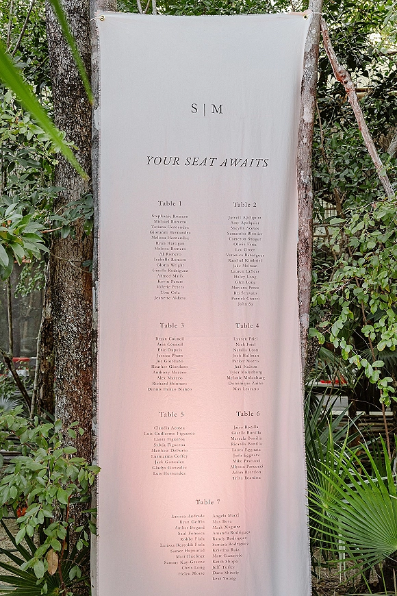 Wedding seating chart fabric banner with printed guest list and grommets, hanging between trees amid lush garden greenery outdoors