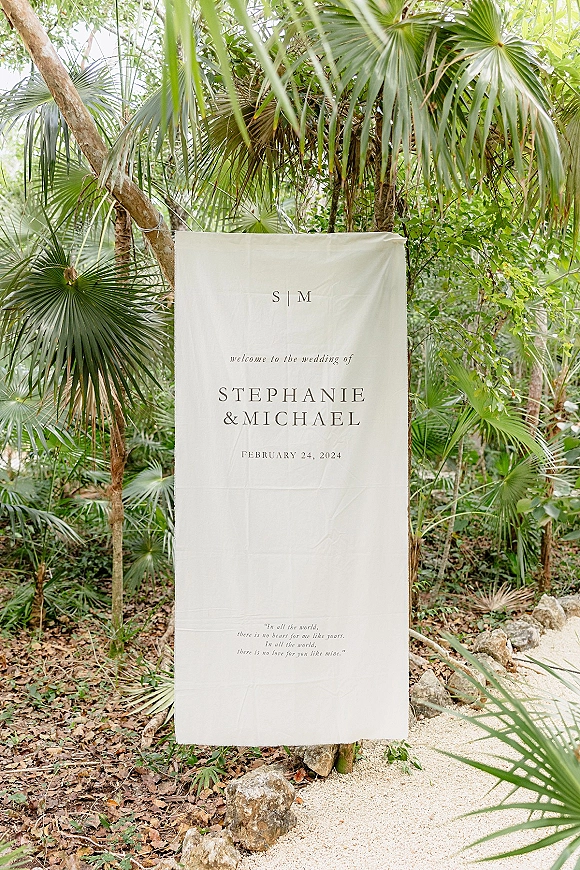 Wedding welcome sign on a linen fabric hanging from a wooden post, displayed among rocks and gravel with tropical palm greenery behind