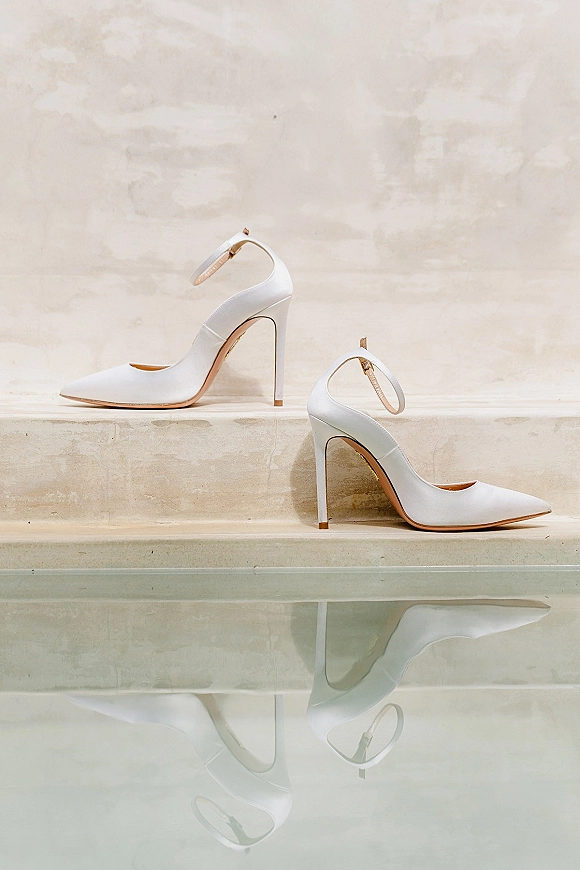 Bridal shoes in white wedding heels with ankle straps resting on stone steps beside a reflective surface against a neutral wall