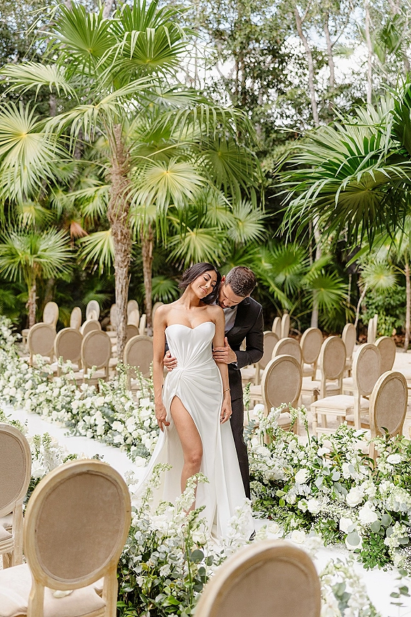 Couple portrait of bride and groom embrace on a floral-lined outdoor ceremony aisle, strapless satin gown and black tuxedo amid palms