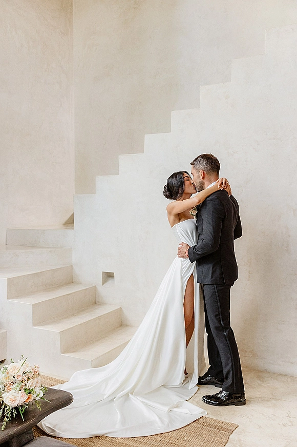 Wedding kiss portrait of bride and groom kissing by a white plaster wall and staircase, her strapless satin dress with long train and slit flowing
