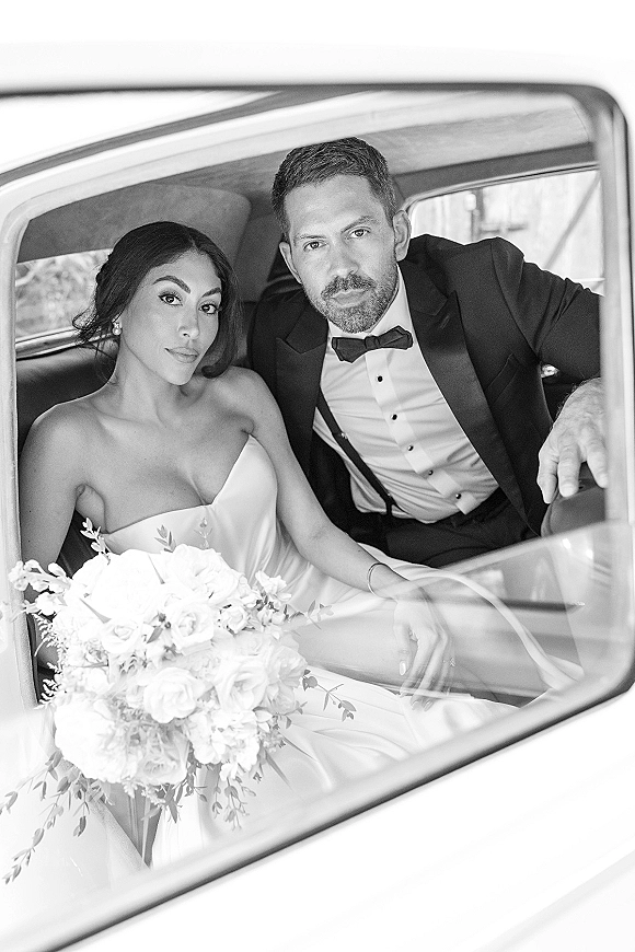 Wedding couple portrait of bride and groom in car, holding a white rose bouquet in a vintage car backseat with blurred scenery outside