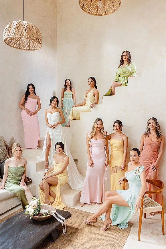 Bridesmaid group portrait with bride and bridesmaids in pastel dresses on an indoor staircase, bouquets in hand, rattan lights above