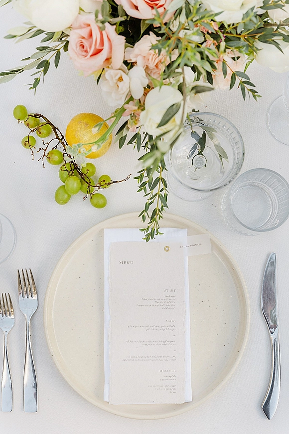 Reception place setting with wedding menu card on a ceramic plate, silver flatware, glassware, and rose centerpiece on a white tablecloth
