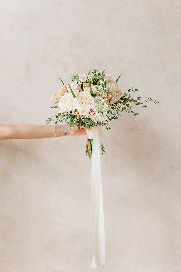 Bridal bouquet of blush roses and ivory roses with greenery and a long white ribbon, held in a bride’s hand against a neutral wall