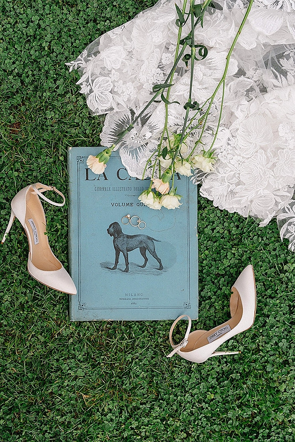 Bridal details flat lay with wedding detail flat lay featuring lace veil, white heels, rings and flowers arranged on clover grass with a vintage book