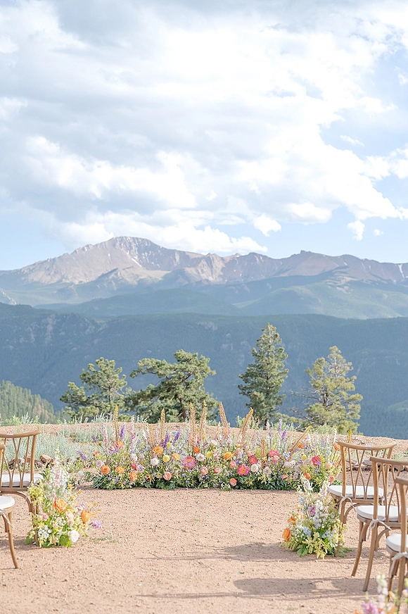 Outdoor ceremony setup with mountain wedding ceremony seating, wooden chairs lining a dirt aisle with pastel floral meadow arrangements under pines