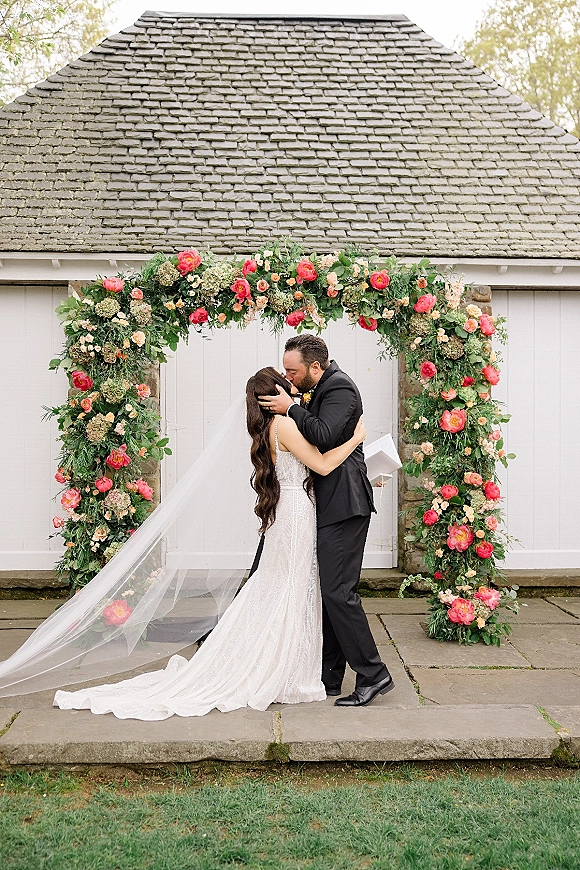 Wedding kiss portrait of bride and groom kissing beneath a lush floral arch, her veil trailing as white doors and stone wall frame them