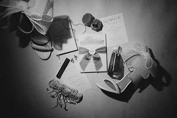 Wedding flat lay with wedding invitation suite, bridal heels, ribbon bow, perfume, lipstick, earrings, garter, and ring box on a plain tabletop