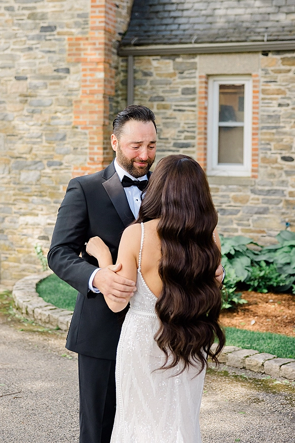 First look moment as groom seeing bride in a beaded wedding gown, embracing on a stone walkway by a brick-and-stone estate garden bed