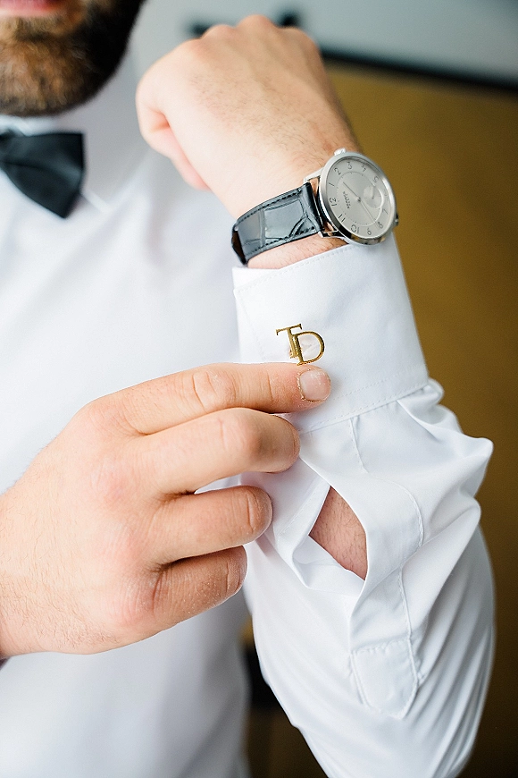 Groom cufflinks with monogram cufflinks in gold on a white dress shirt, paired with a wristwatch and black bow tie in soft indoor light