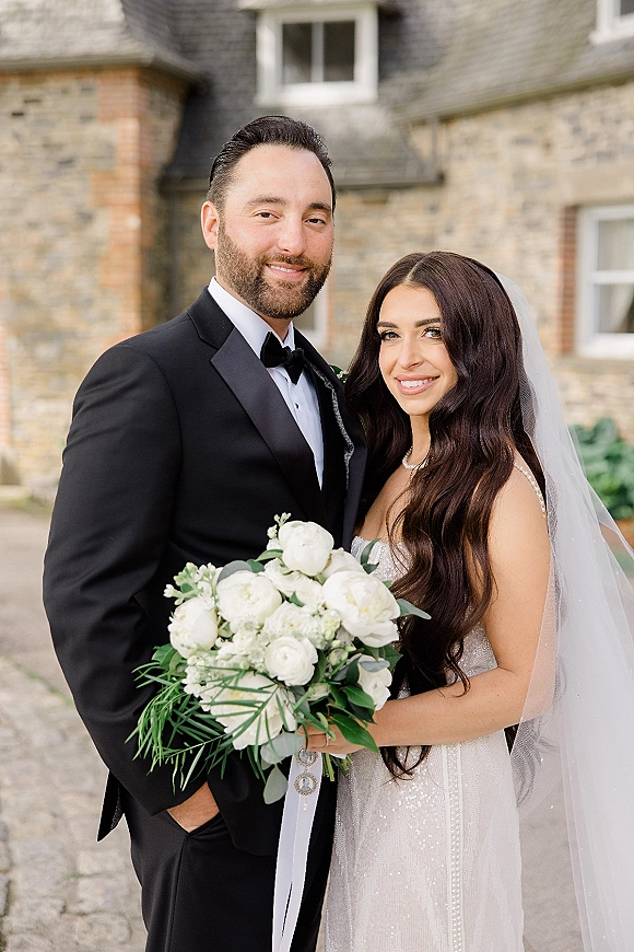 Couple portrait of bride and groom smiling, bride holding white rose bouquet with greenery and veil before a stone building courtyard